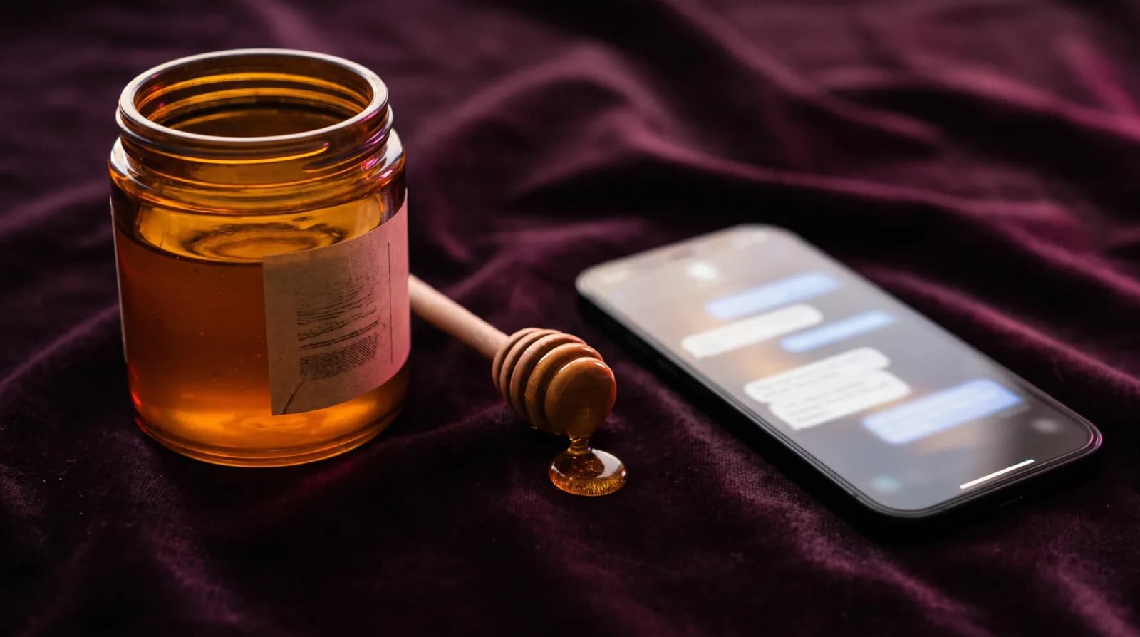 A jar of honey with a wooden dipper beside a smartphone showing blurred chat bubbles on dark plum velvet